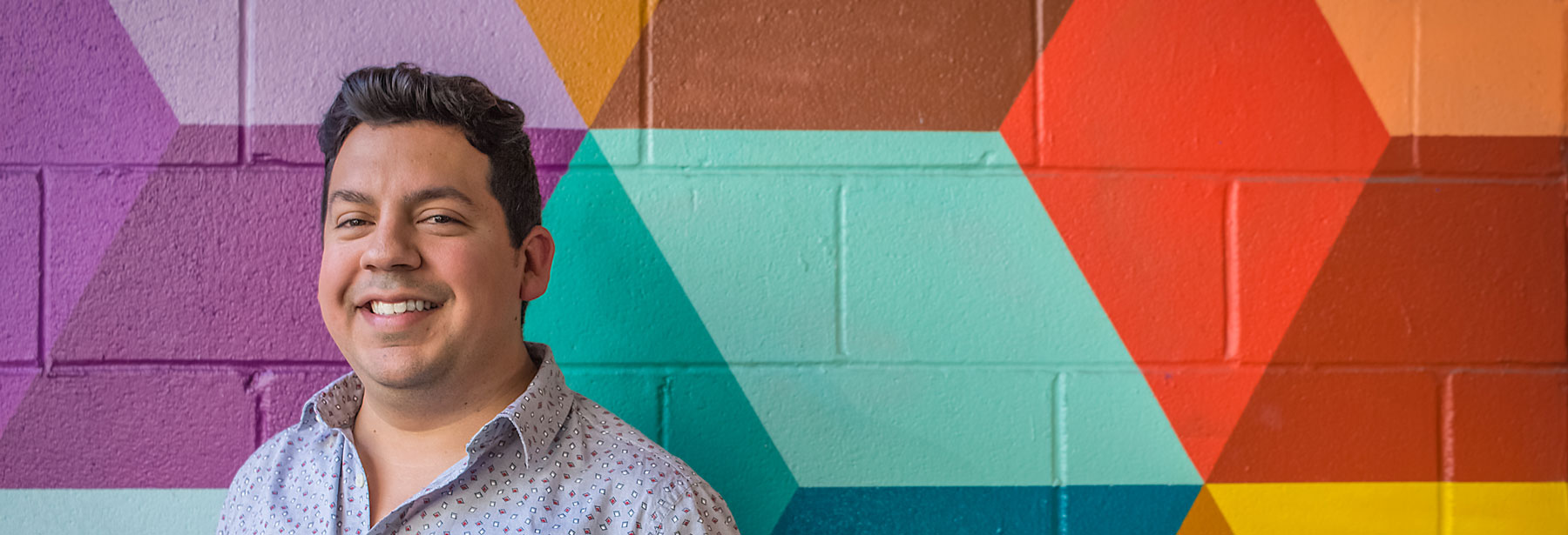 round Latino man smiling at the camera against a multi-colored hexagon background