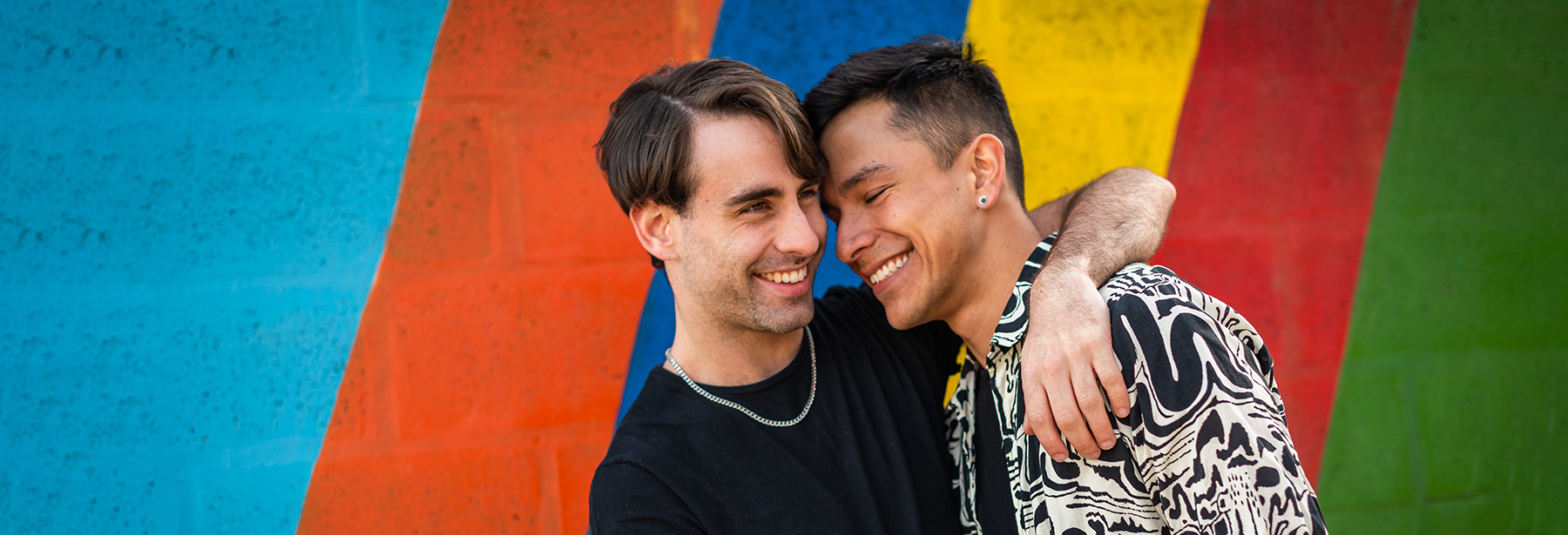 Two young men embracing in front of a colorful, striped wall.