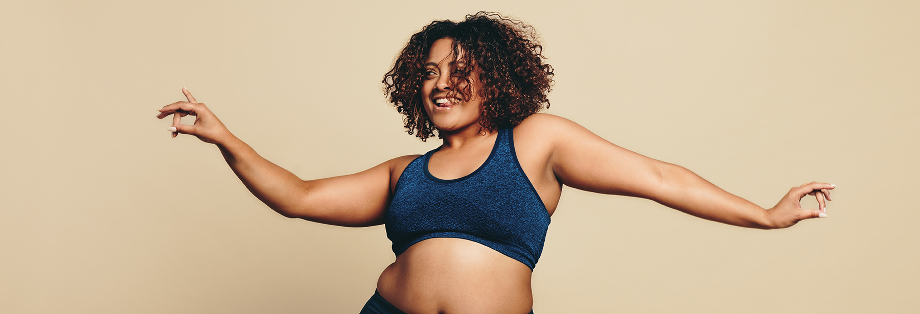 Woman with curly hair in workout clothes dancing against a plain, tan background.